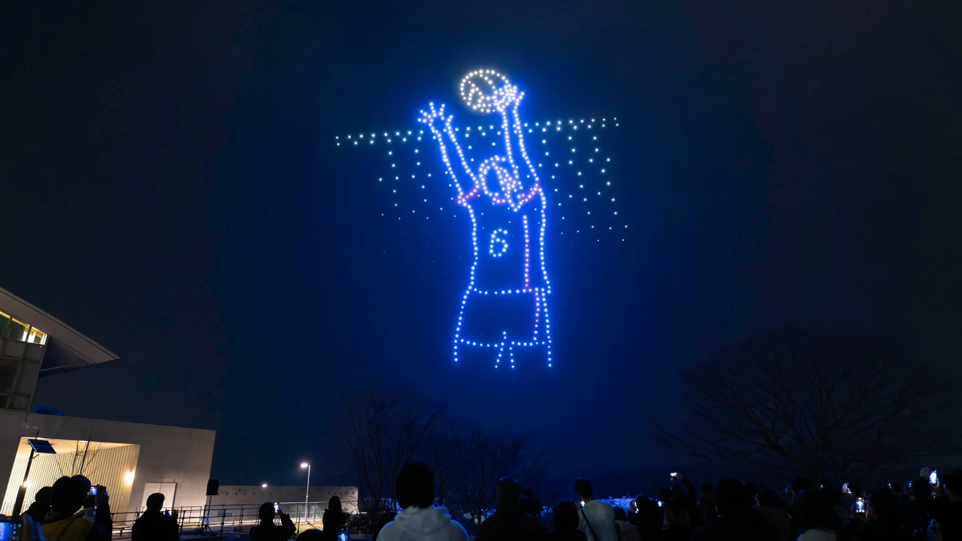 夜空に多数のドローンが青い光でバレーボール選手を形作り、スパイクを打つ瞬間を表現