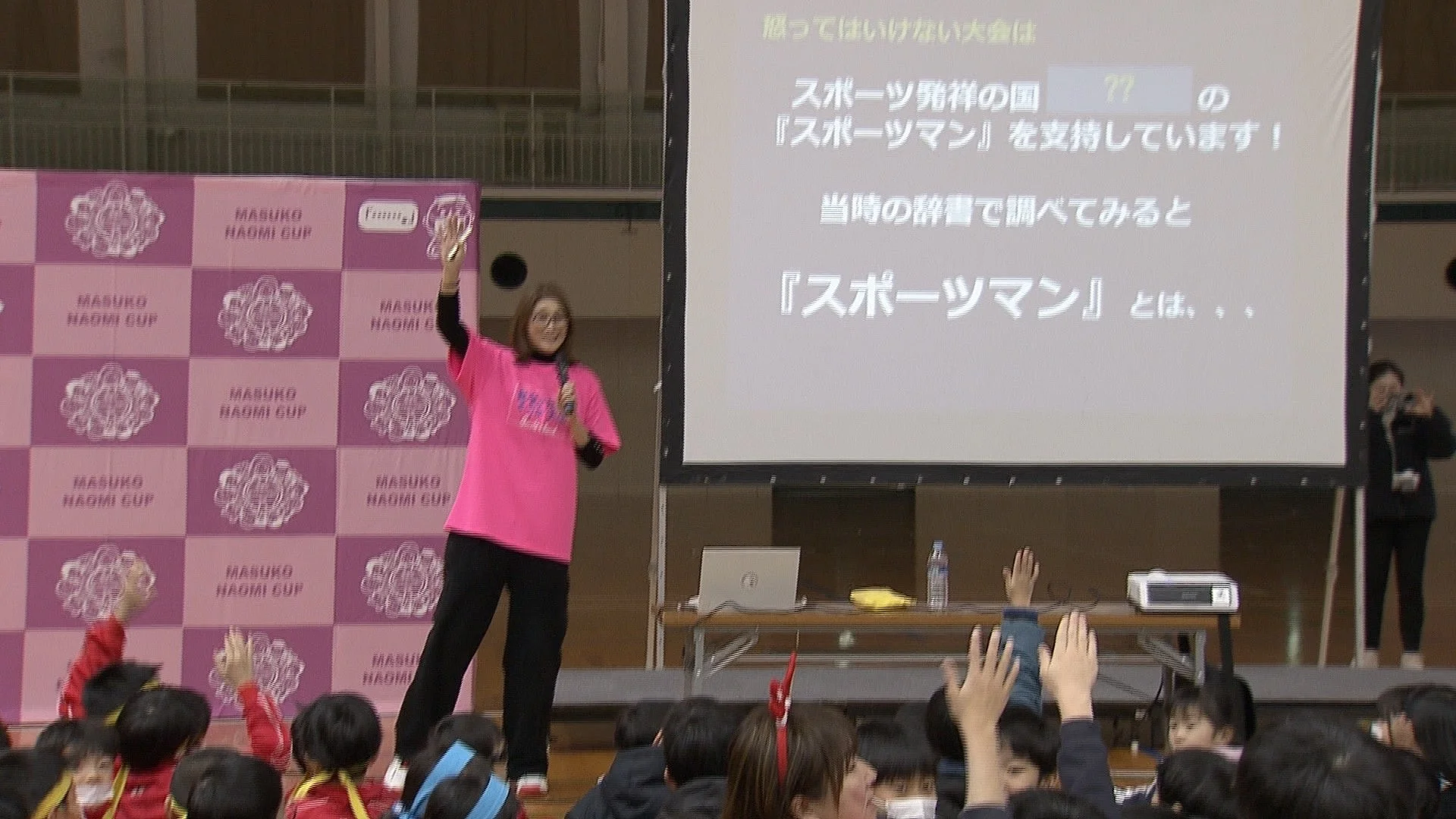 ピンクのシャツを着た女性が、ステージ上で子供たちにプレゼンテーションを行っています