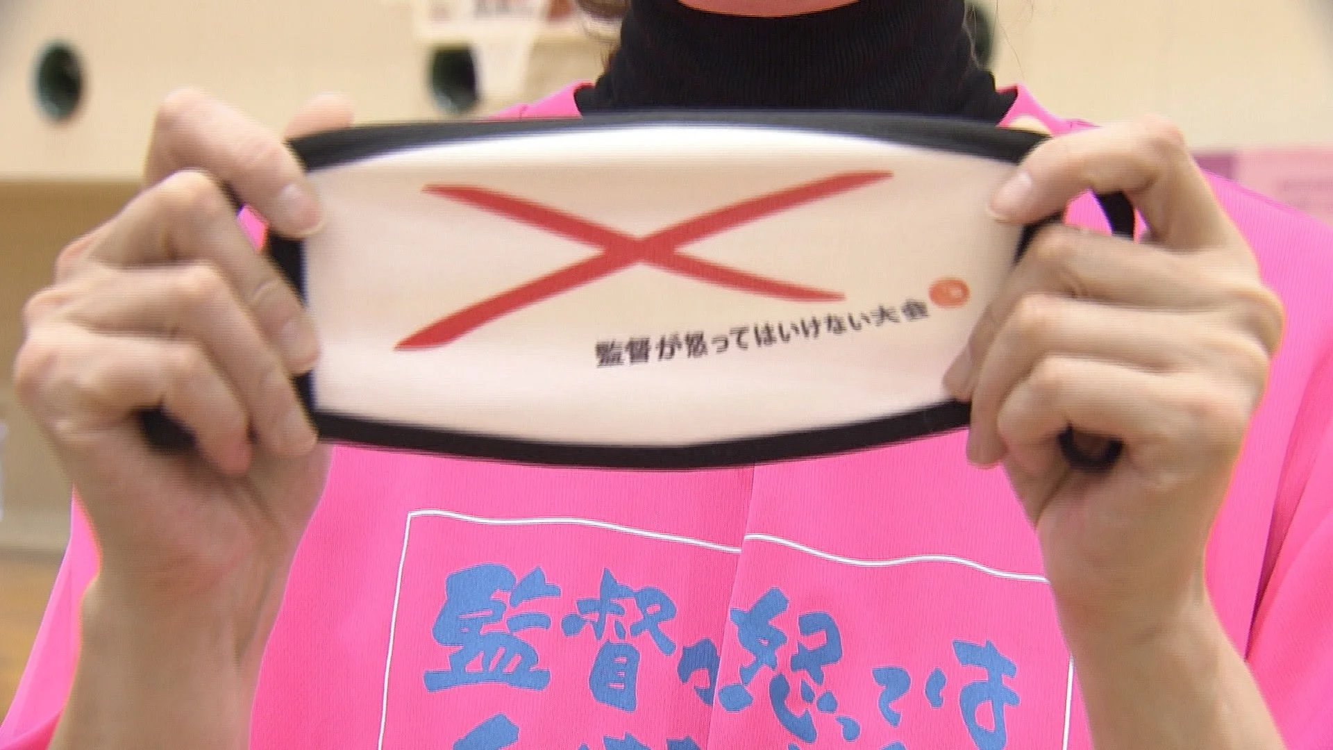 ピンクのTシャツを着た人物が「監督が怒ってはいけない大会」と書かれた白い布（マスクと推測される）を掲げている