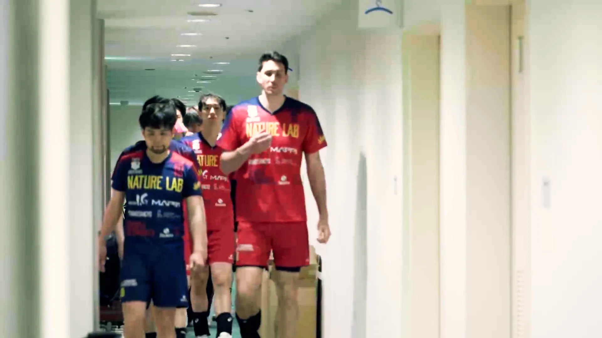 チームのユニフォームを着た複数の男性選手が、明るい屋内の通路を歩いている様子