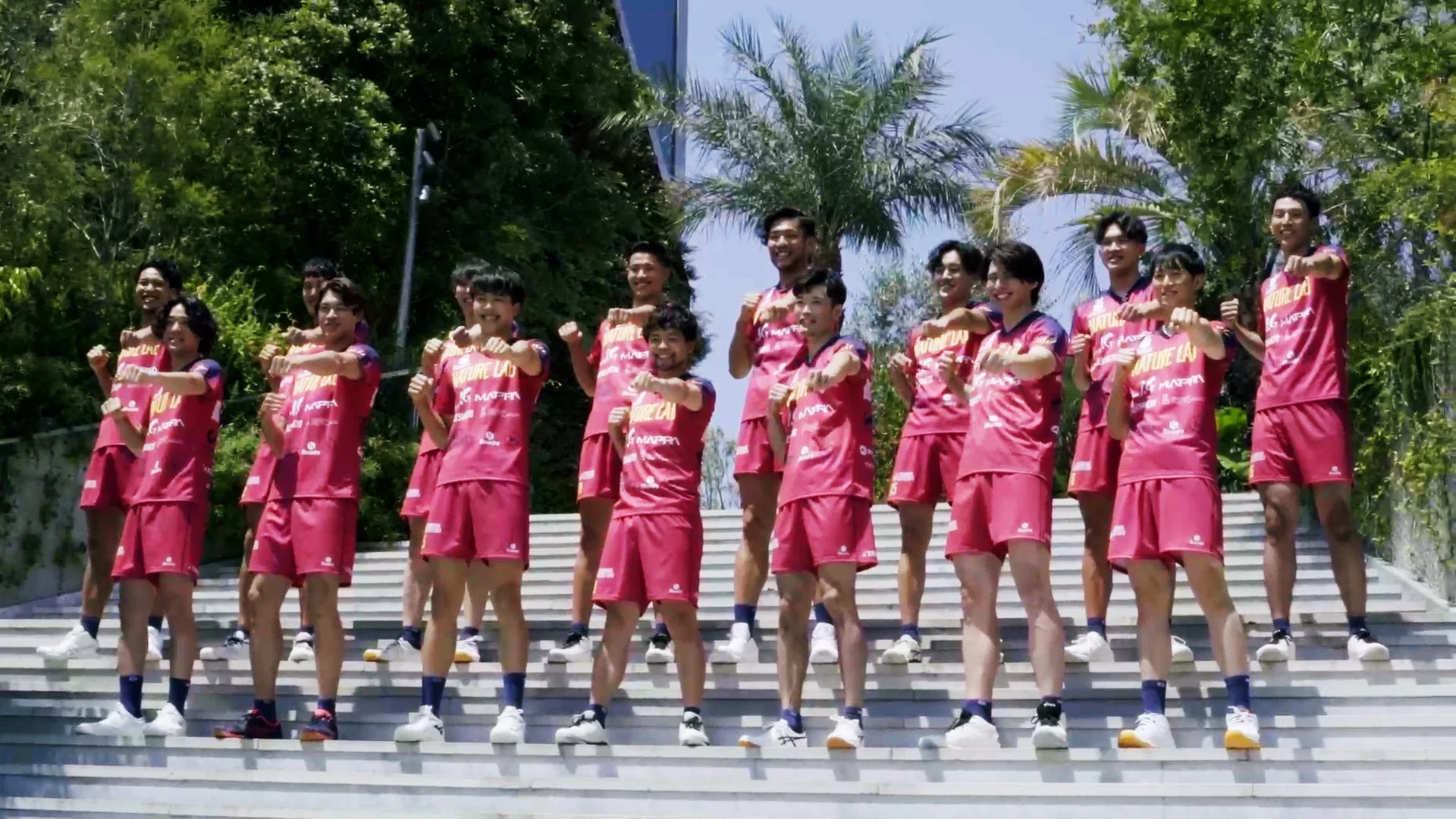 屋外の階段に立つ、ピンク色のユニフォームを着た男性のスポーツチームが、皆で同じポーズをとっている
