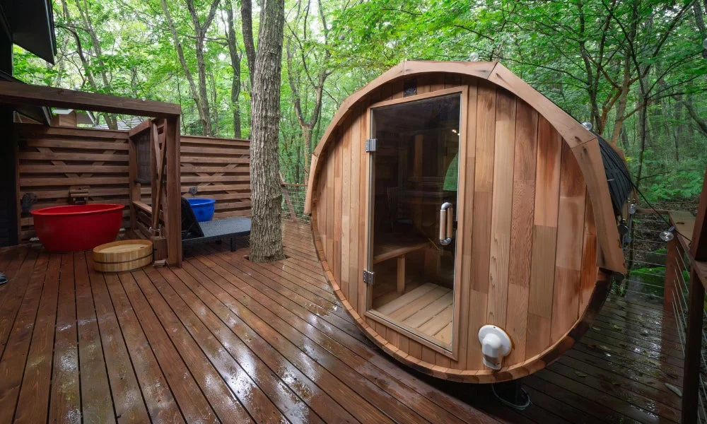 森林に囲まれたウッドデッキに、樽型のバレルサウナと赤い浴槽、木桶が配置されたアウトドアのリラックス空間です。