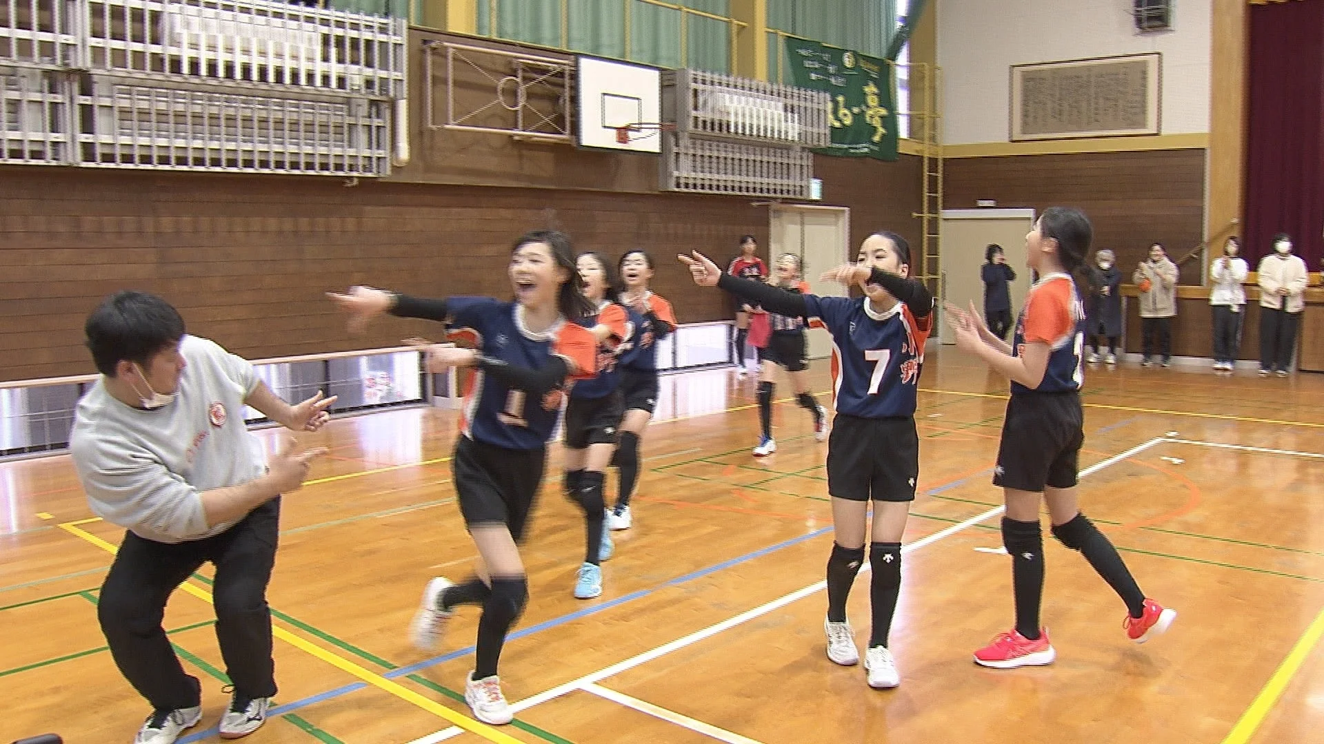 体育館で、女子学生たちがバレーボールのユニフォームを着て、コーチらしき男性と共に活気ある練習やゲームを楽しんでいる様子