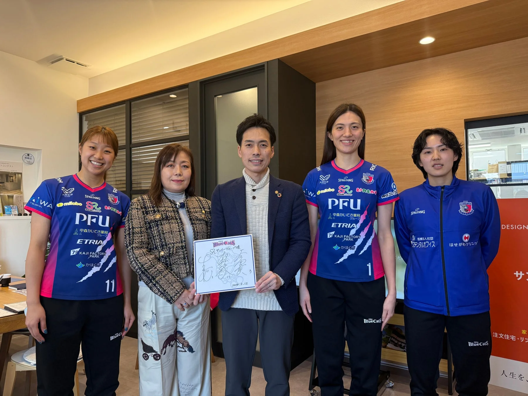 PFUブルーキャッツのユニフォームを着用したバレーボール選手3名を含む5名が、室内で記念撮影をしている様子