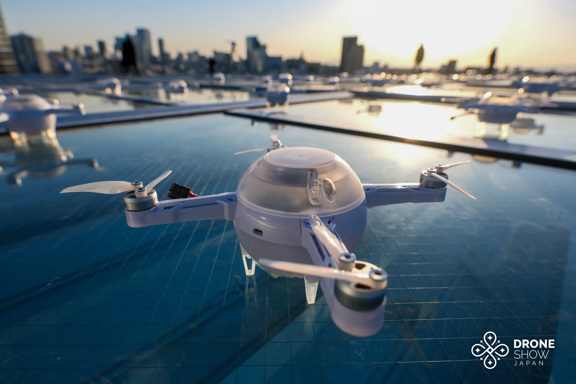 夕焼け空を背景に、反射する床の上に置かれた球状の白いドローンが複数並んでいます