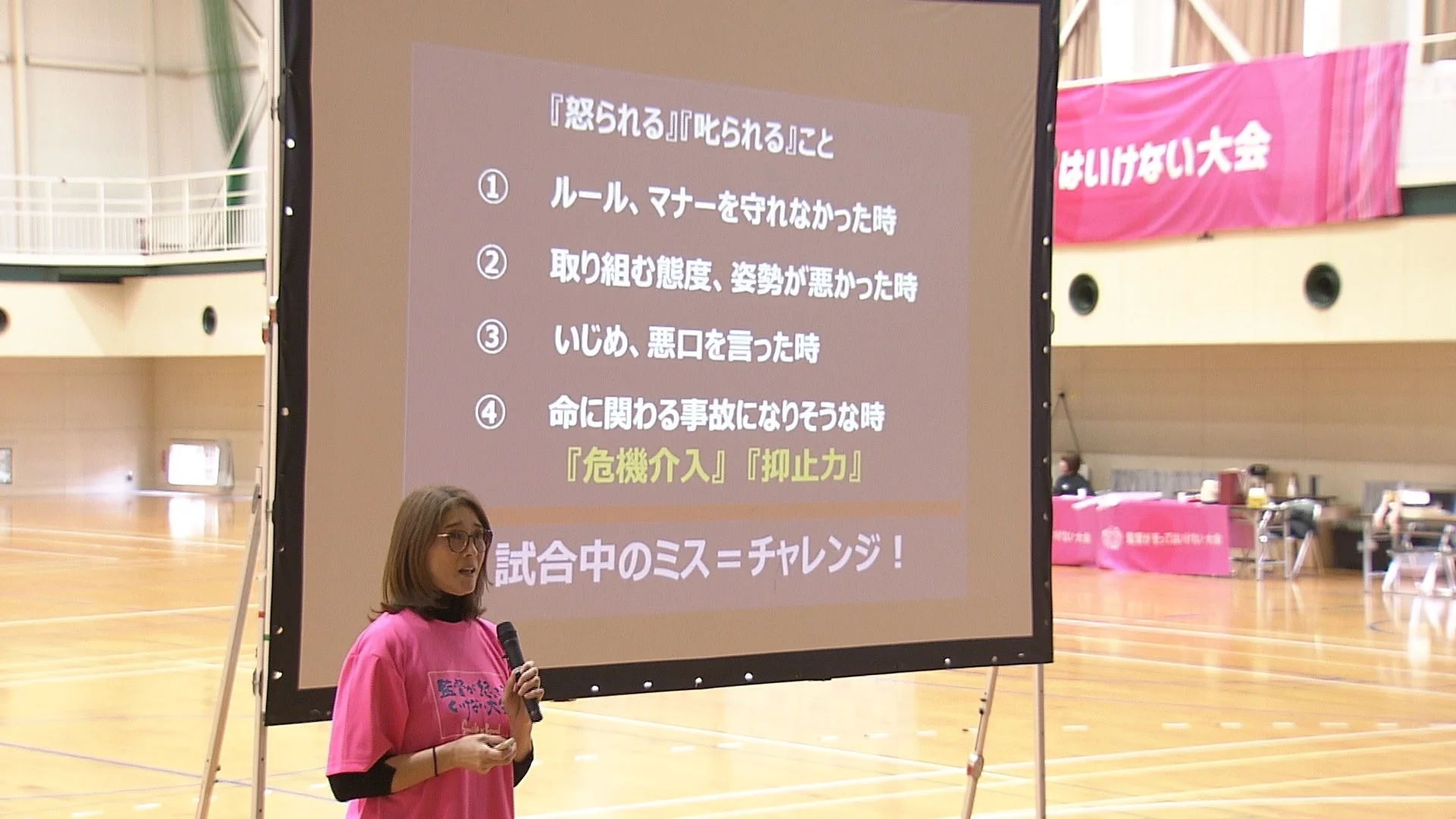 体育館で女性が講演しており、プロジェクターには「怒られる」「叱られる」状況として、ルール違反、態度、いじめ、危険回避の重要性が示されている