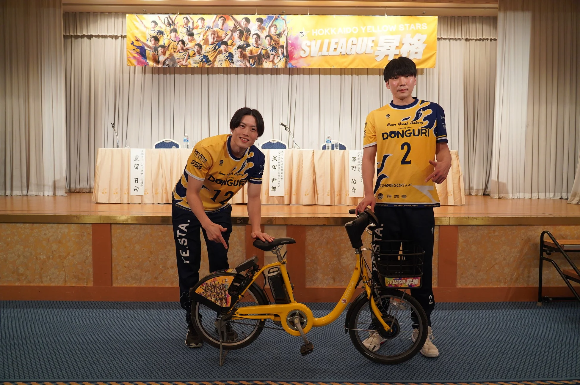 北海道イエロースターズの選手2名がSVリーグ昇格イベントで、スポンサーDONGURIのユニフォームを着用し、電動アシスト自転車と共に笑顔でポーズ。背景には昇格バナー。