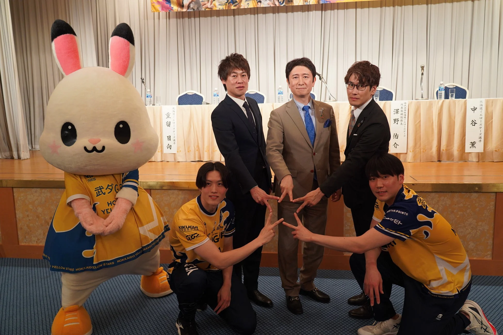 うさぎのマスコットキャラクターと、スーツ姿の男性3名、黄色と青のユニフォーム姿の選手2名が星のポーズをとる集合写真。記者会見のような会場で、北海道イエローロースターズ関連のイベントと見られる。