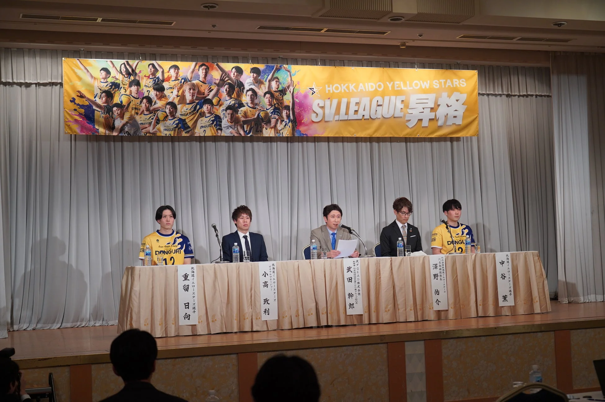 バレーボールチーム「北海道イエロースターズ」のSV.LEAGUE昇格に関する記者会見の様子です。選手や関係者らが壇上に並び、発表を行っています。