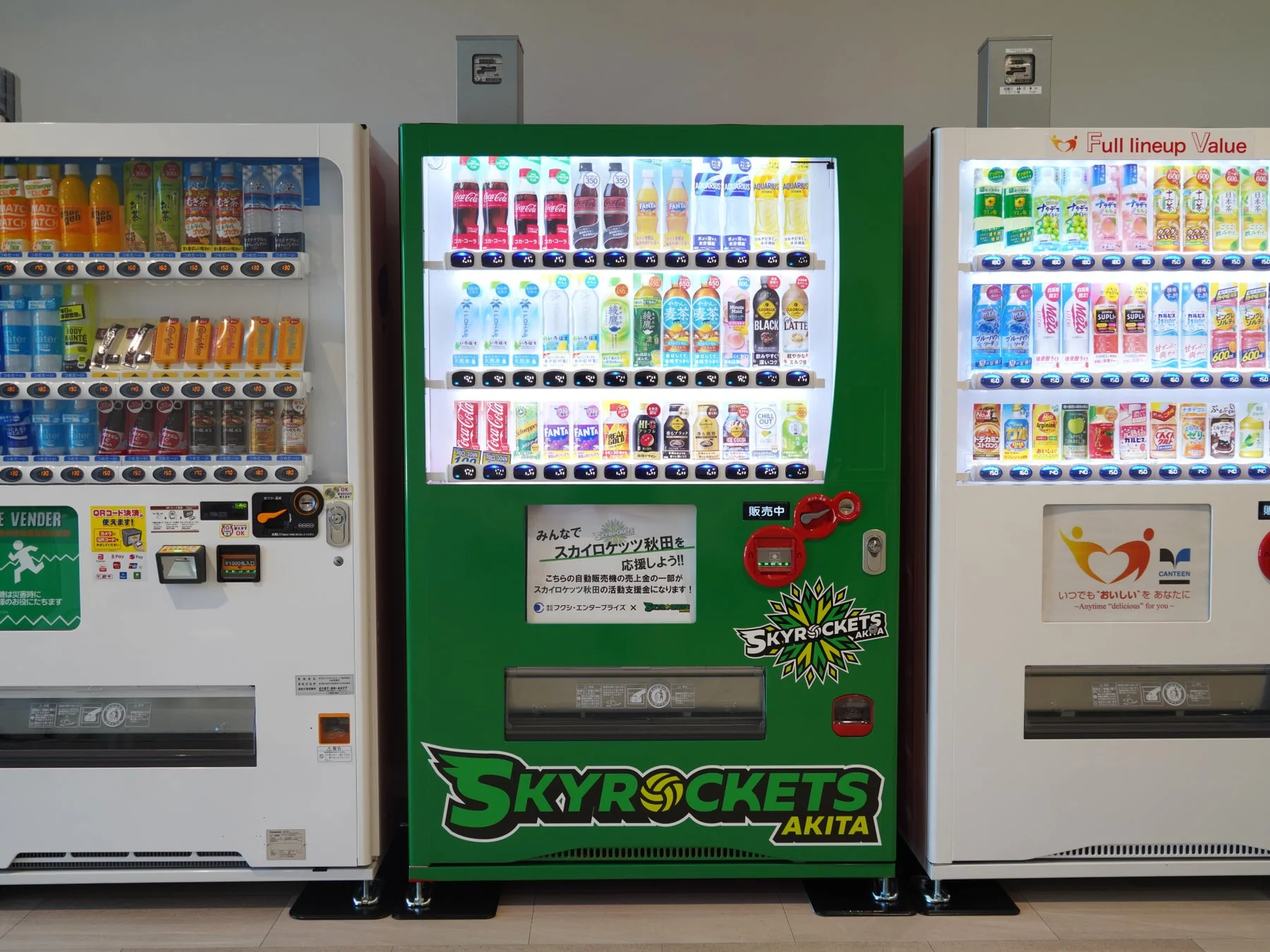 3台の自動販売機が並ぶ室内の様子。中央の緑色の自販機は「SKYROCKETS AKITA」のロゴがあり、売上の一部が活動支援金になるチャリティー自販機。