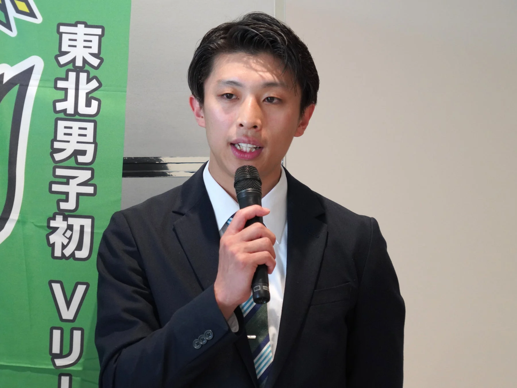 スーツを着た男性がマイクを持ってスピーチしている様子。背景には「東北男子初Vリ」と書かれた緑色のバナー