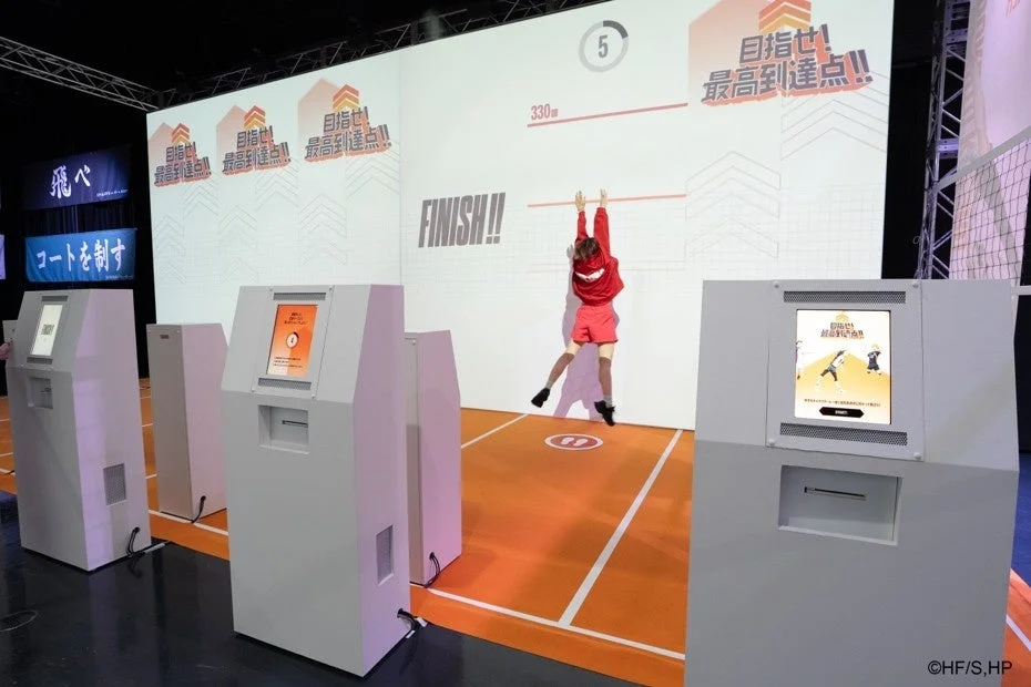 バレーボールをテーマにした体験型アトラクションで、参加者がスクリーンに向かってジャンプし、最高到達点を目指している様子です。画面には「目指せ! 最高到達点!!」や「FINISH!!」などの文字が表示され、複数の受付らしきキオスクが設置されています。