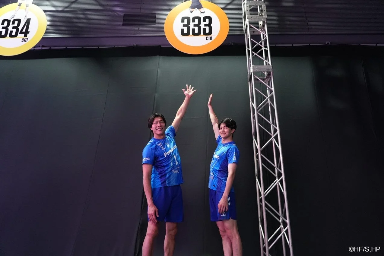 青いユニフォームを着た2人の男子バレーボール選手が、天井の高さを示すマークに向かって手を伸ばし、笑顔でポーズをとっている。
