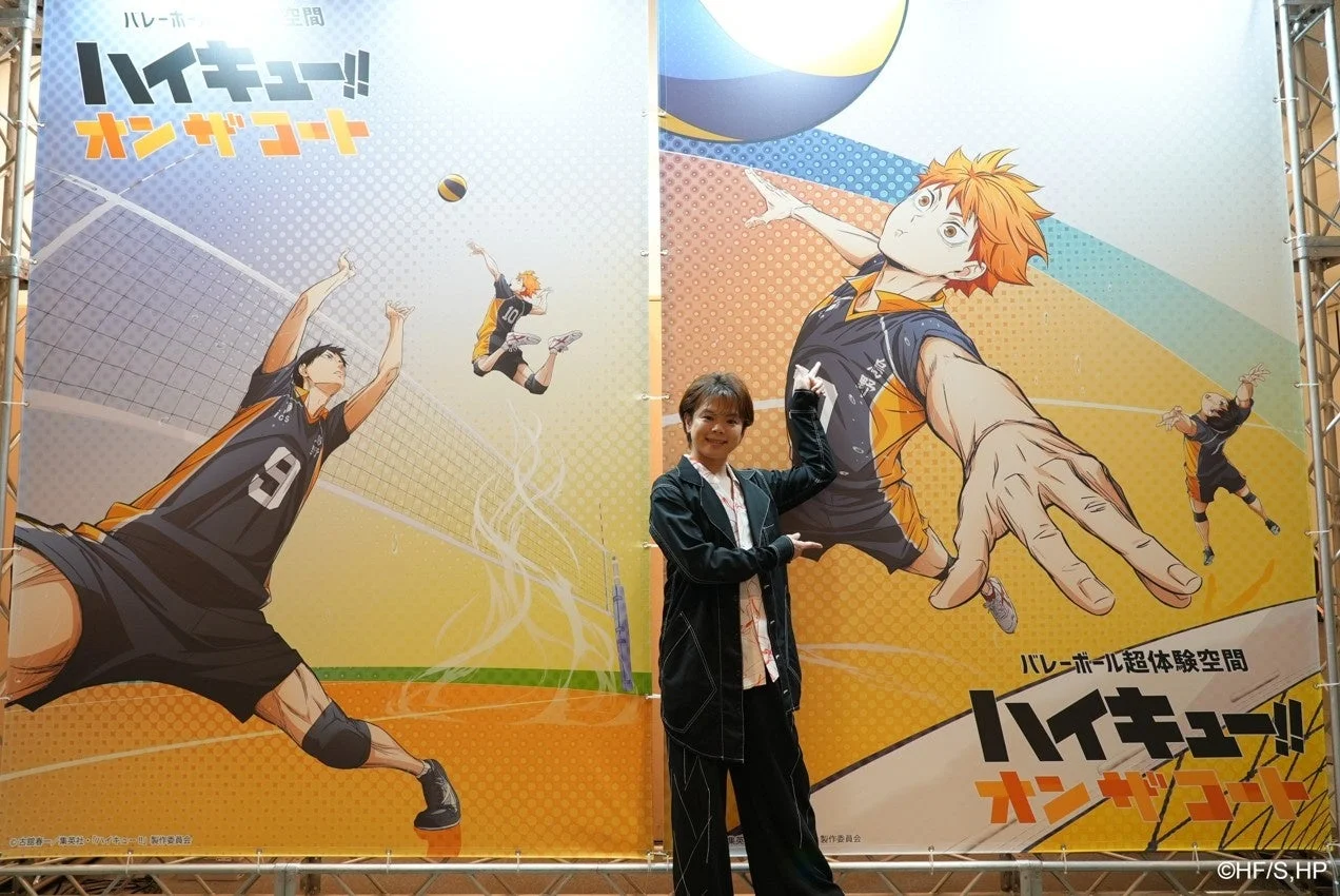 アニメ『ハイキュー!!』の大型バナーの前でポーズを取る人物。バナーにはバレーボールをする日向翔陽や影山飛雄のイラストが描かれ、「ハイキュー!! オンザコート」というイベント名が見える。バレーボールの超体験空間をPRしている様子。