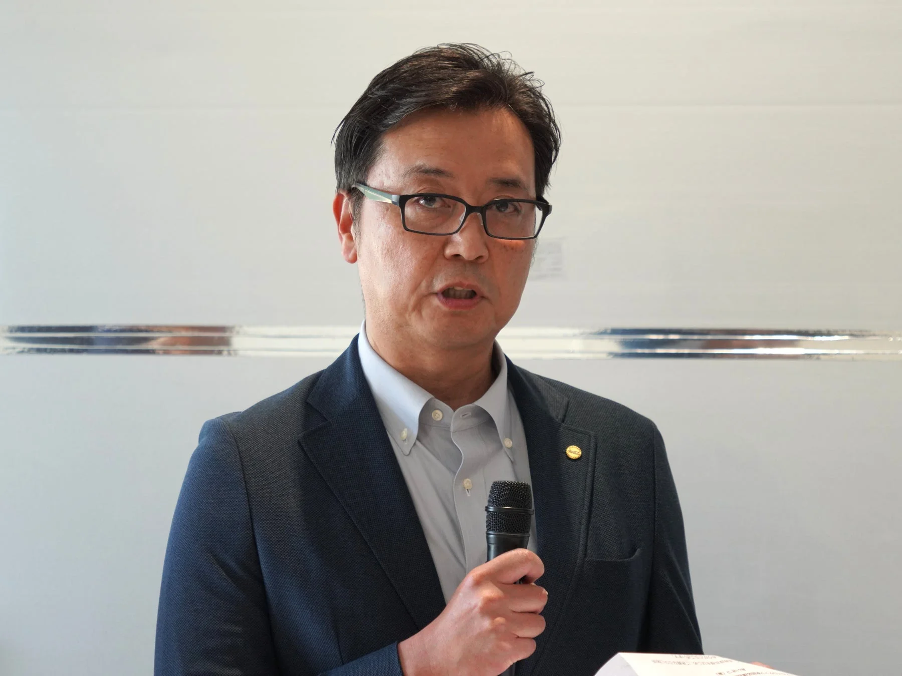 スーツと眼鏡を着用した東洋人男性がマイクを持ち、何かを話している様子