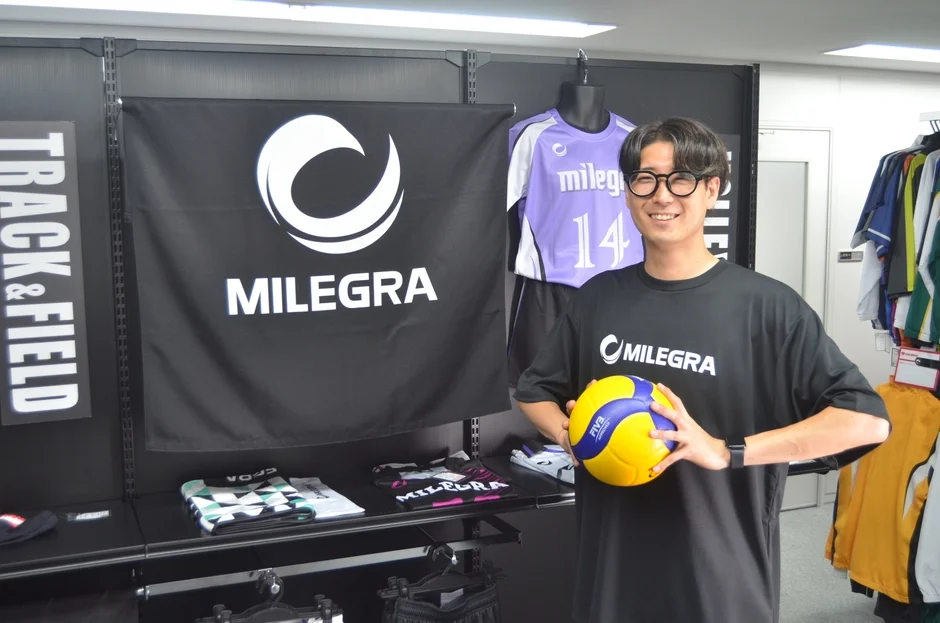 スポーツウェアの店舗で、MILEGRAのTシャツを着た男性が笑顔でバレーボールを抱えている。背景にはMILEGRAのロゴ入りバナーやユニフォームが展示されており、スポーツ用品店の様子がわかる。