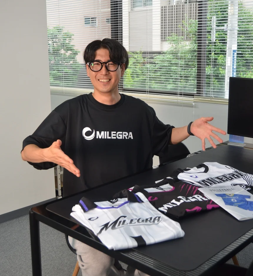 MILEGRAのTシャツを着た男性が、デスクに並べられたMILEGRAブランドのスポーツウェア(ユニフォーム)を笑顔で紹介しています。