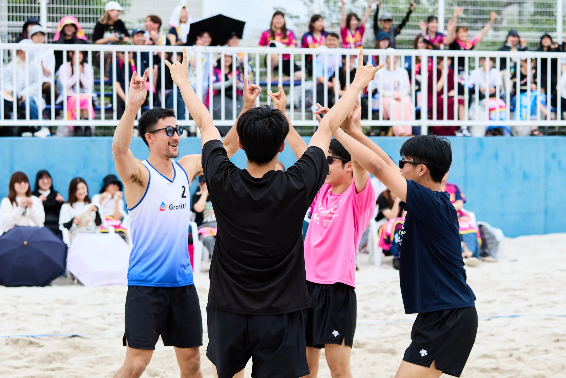 ビーチバレーの試合中、4人の男性選手が両手を上げて喜びを分かち合っている