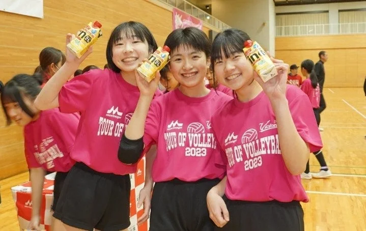 体育館でピンクのバレーボールユニフォームを着た3人の女子学生が、笑顔でプロテインドリンクを持っている様子です。スポーツイベントの一コマのようです。