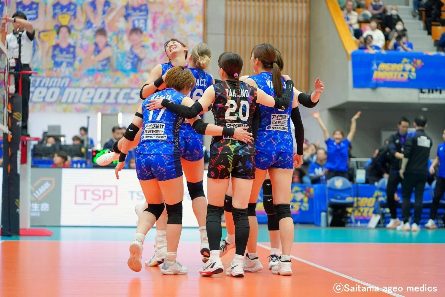 バレーボールの試合で、青いユニフォームを着た女子選手たちが円陣を組み、勝利や好プレーを喜んでいる瞬間です。選手たちは笑顔で互いを抱きしめ合い、強いチームワークが感じられます。