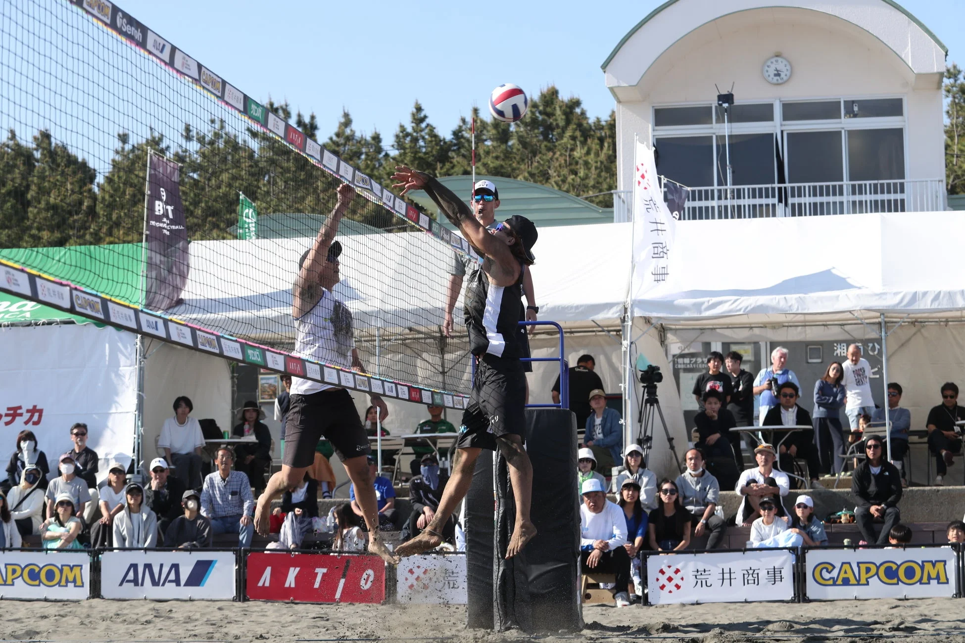 青空の下、ビーチバレーの試合が行われている様子。2人の選手がネット際でボールを打ち合っており、多くの観客が試合を観戦している。