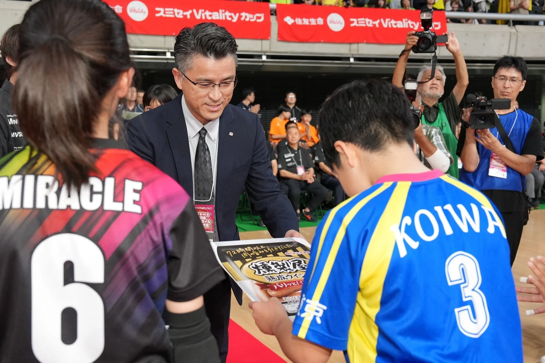 屋内スポーツイベントの表彰式で、スーツ姿の男性がユニフォームを着た選手たちに賞状のようなものを手渡している場面